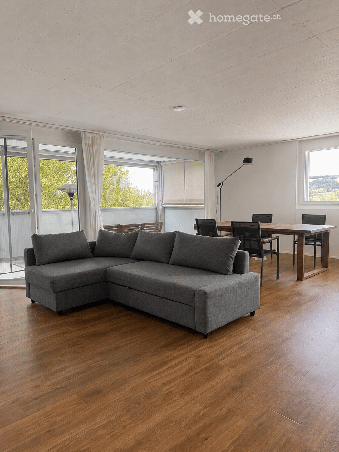 Modern living and dining area with a grey sectional sofa, wooden dining table, and chairs, large windows with curtains, and a view of greenery outside.