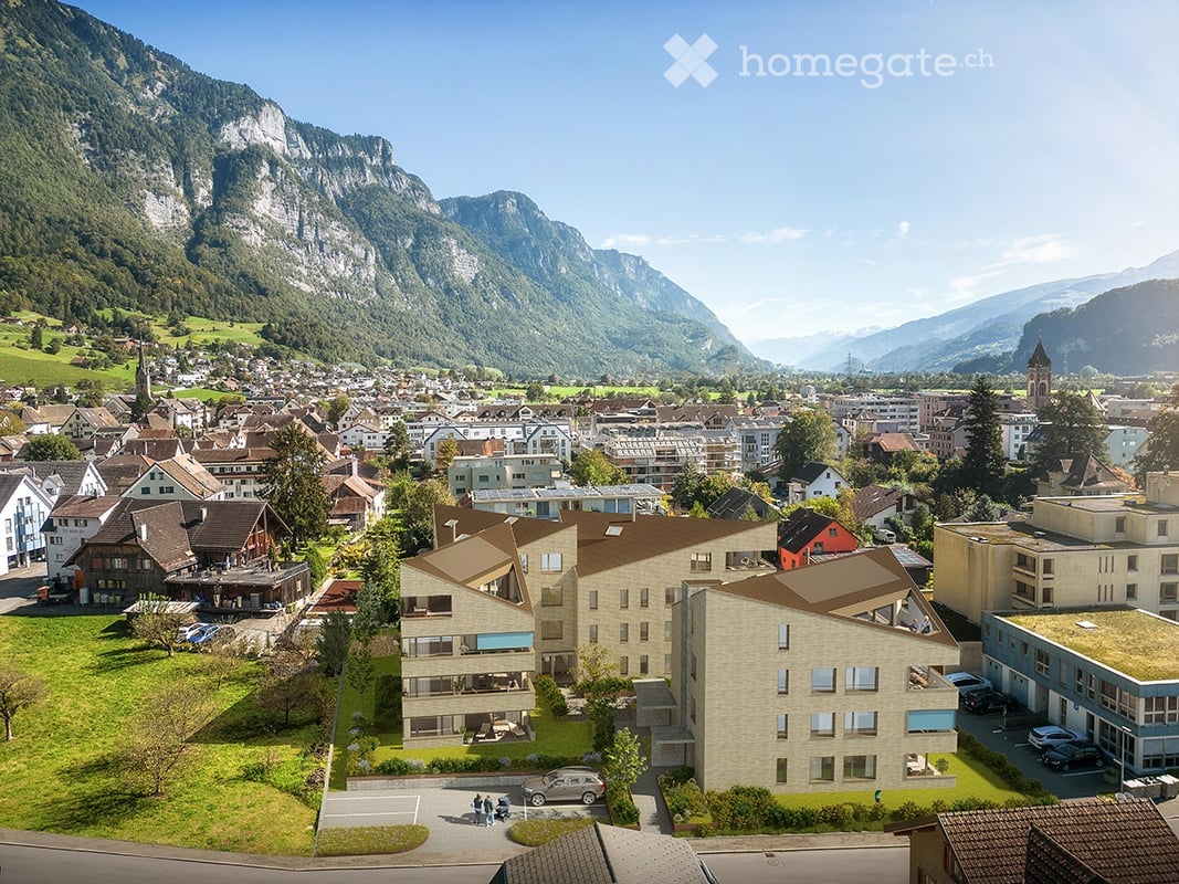 A modern multi-story building with a contemporary design, situated in a picturesque town surrounded by lush green hills and mountains.