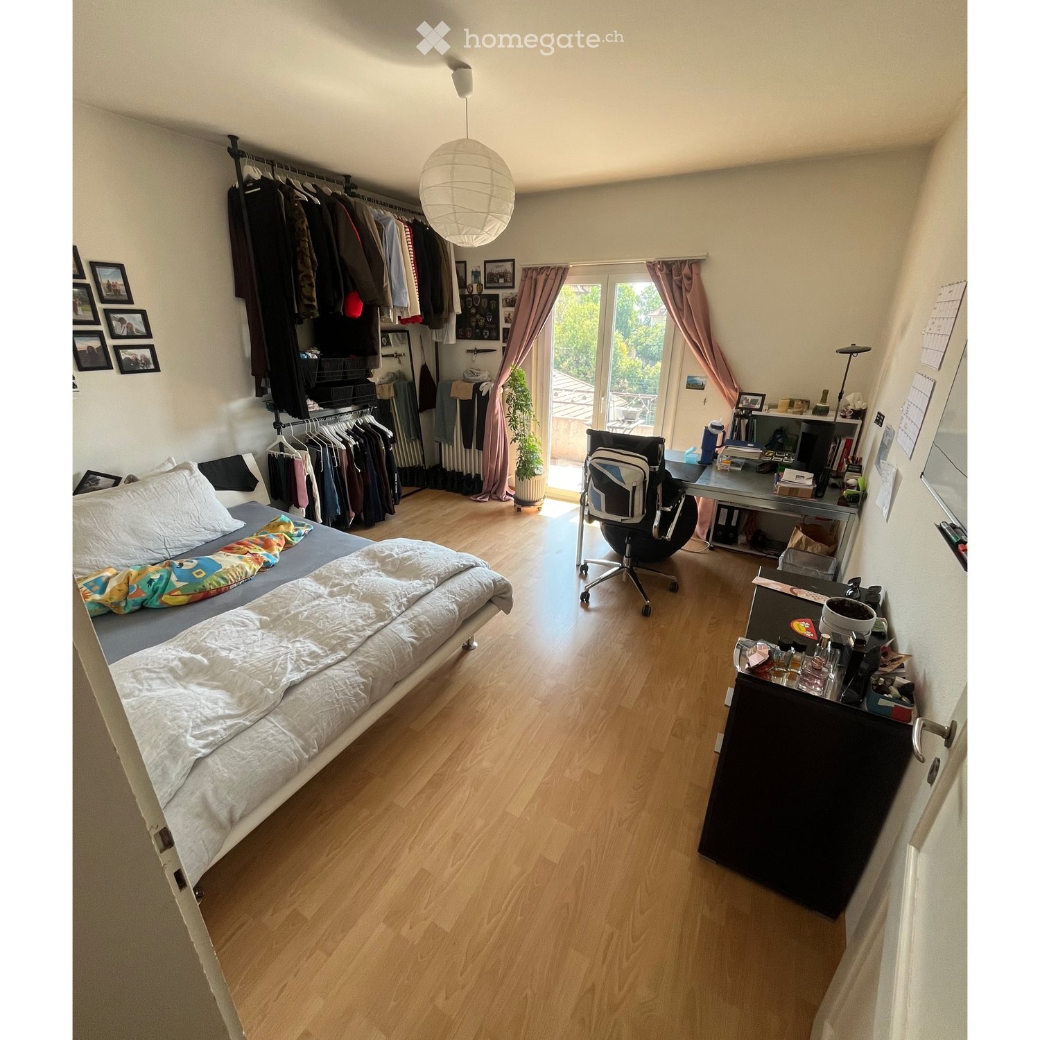 A well-lit bedroom with a bed, desk, chair, and a small closet. The room features wooden flooring, a large window with pink curtains, and a hanging pendant light.