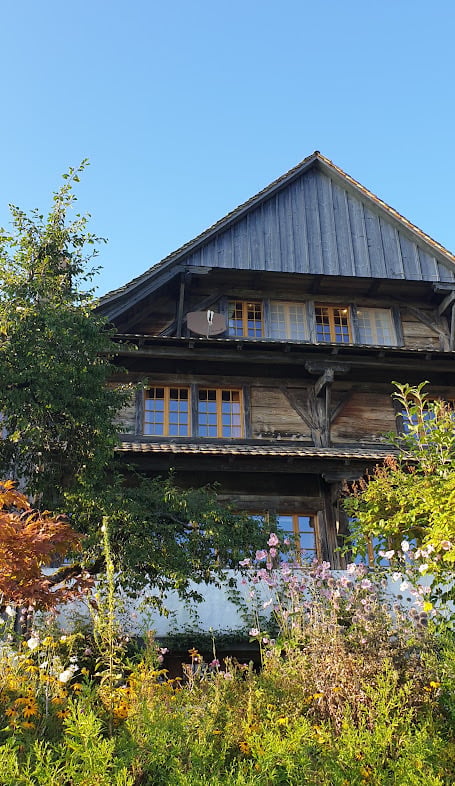 A two-story wooden house with a steeply pitched roof, large windows, and a balcony on the upper floor, surrounded by lush greenery and flowers.