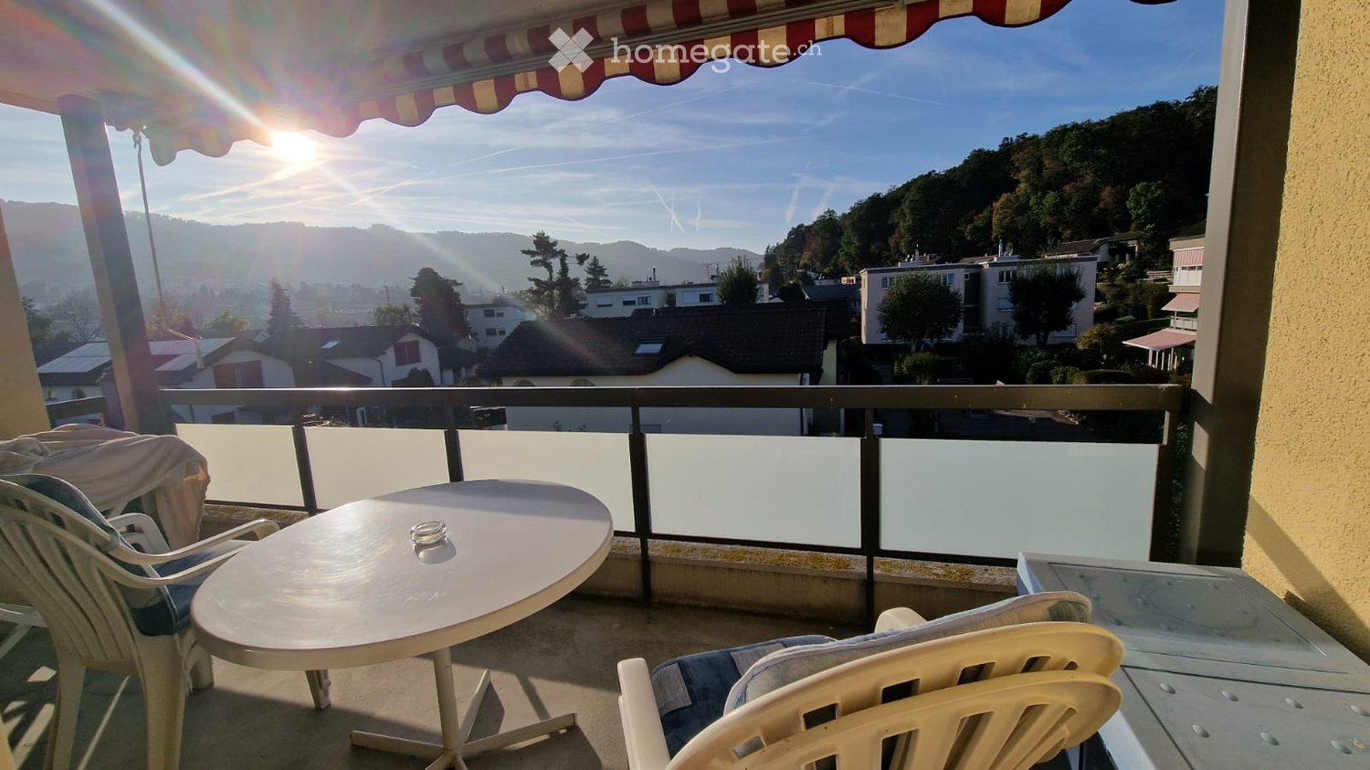 Balcony with a table and chairs, overlooking a scenic town and hills under a clear sky.