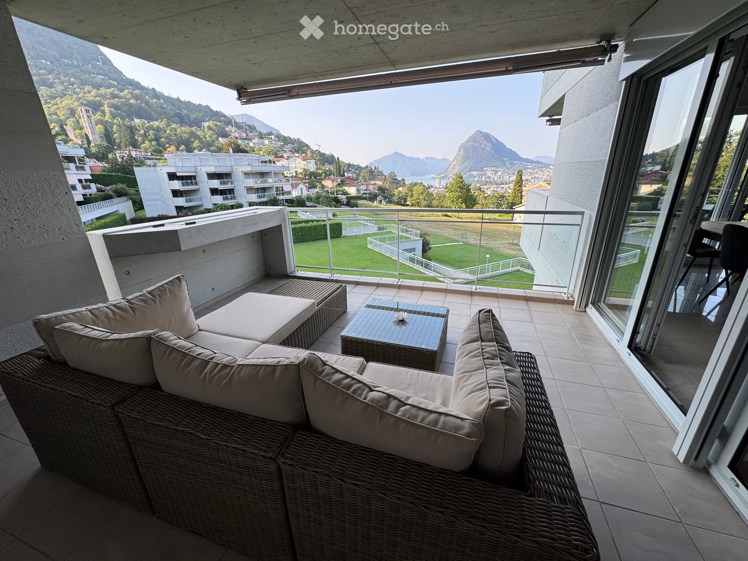 Balcony with a view of a cityscape and mountains, featuring a wicker sofa, coffee table, and chair.