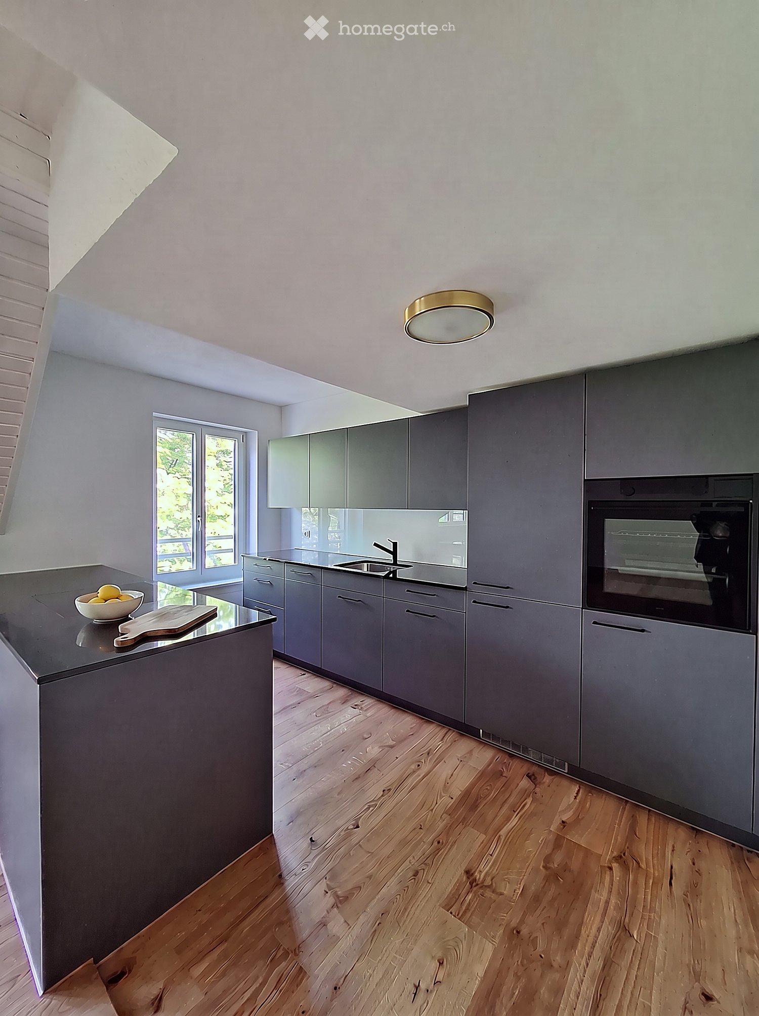 Modern kitchen with grey cabinets, wooden flooring, island, sink, and oven. Features a bowl of fruit and a cutting board on the island.
