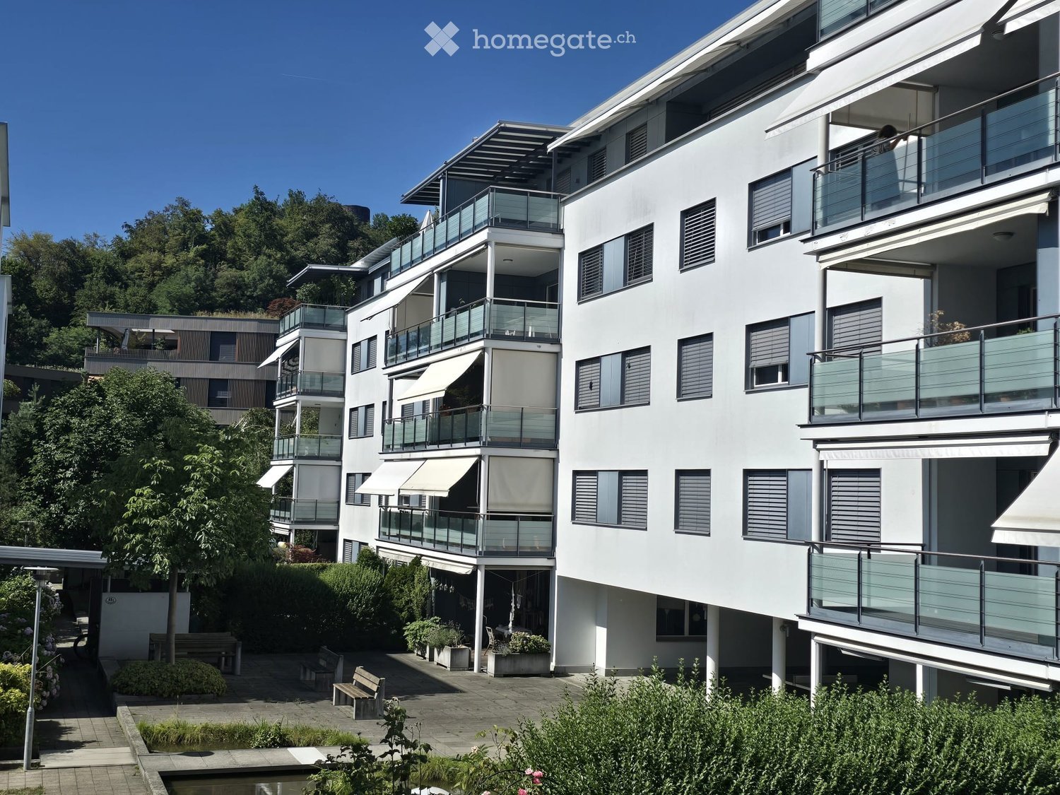 Modern multi-story residential building with white facade, large windows, and multiple balconies. The building features contemporary design with glass railings and awnings. The surrounding area includes landscaped gardens, benches, and a small water featu