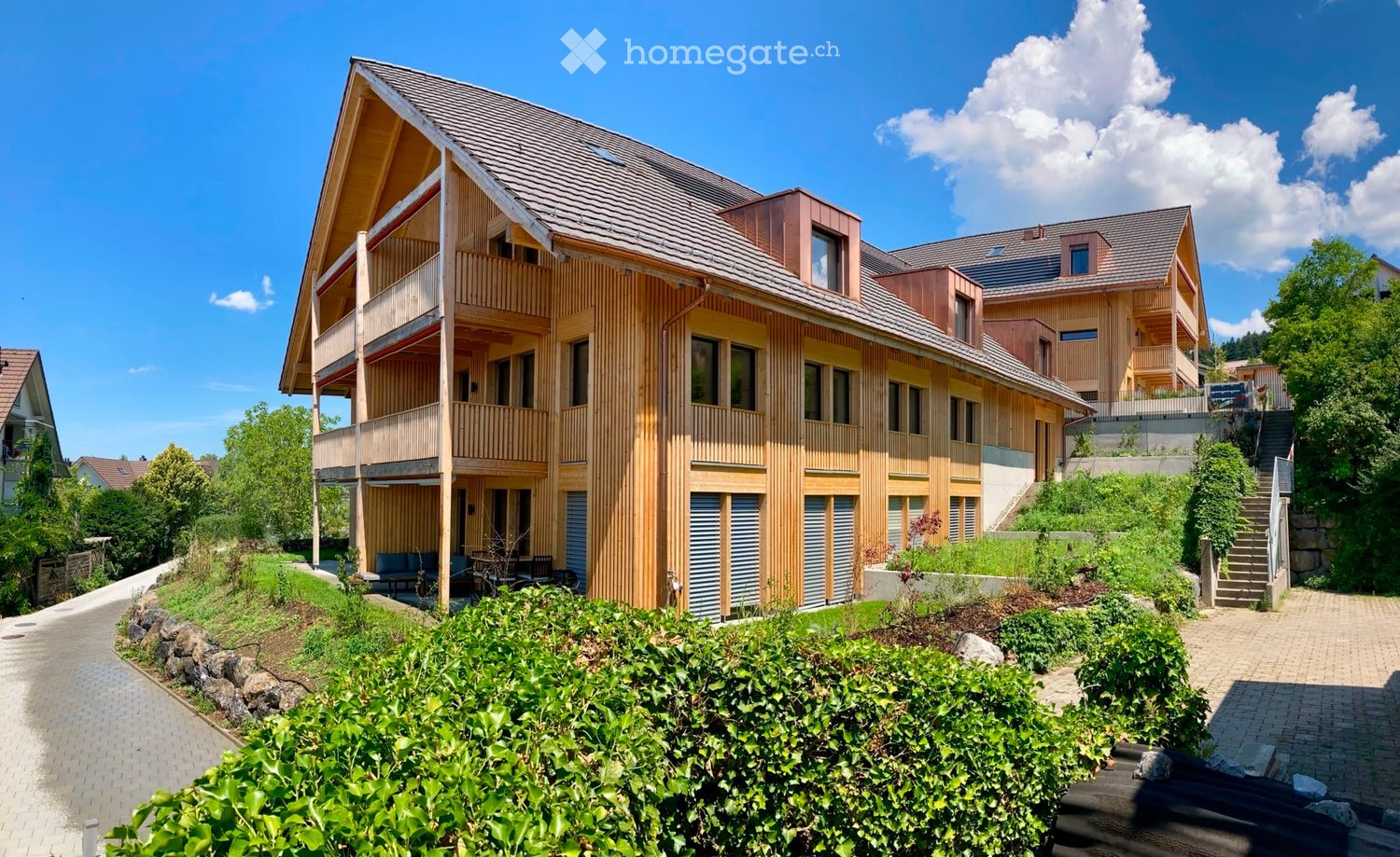 Modern two-story residential building with wooden facade, large windows, and balconies. The building is surrounded by greenery and has a paved pathway leading to the entrance.