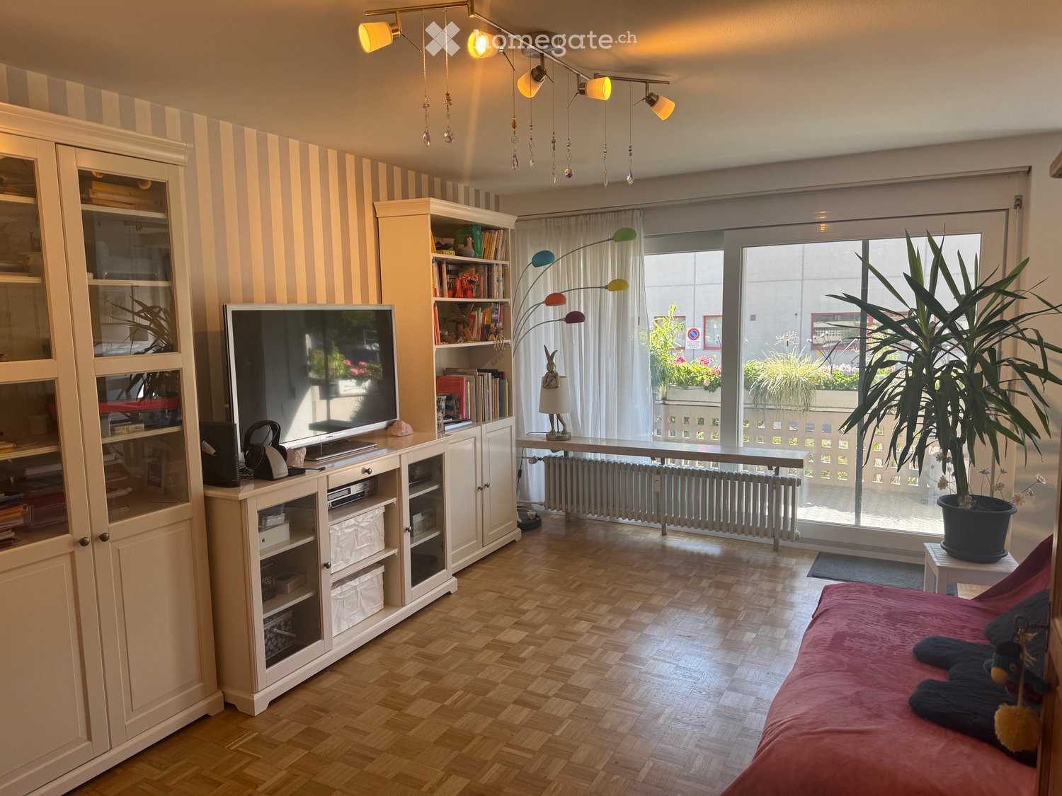 Bright bedroom with wooden flooring, large window with white curtains, bed with pink bedding, cabinet, bookshelf, table with lamp, and potted plant.