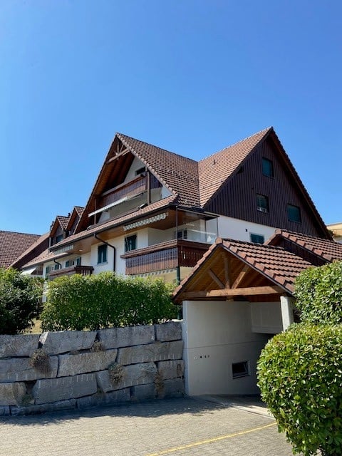 Two-story house with a red-tiled roof, wooden facade, and a garage entrance. The house features balconies and is surrounded by greenery.