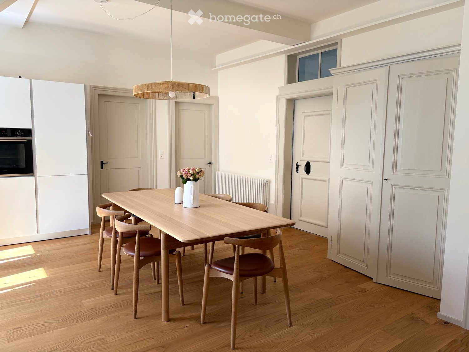 Modern dining area with a wooden dining table, four chairs, a vase with flowers, and built-in white cabinets with an oven.