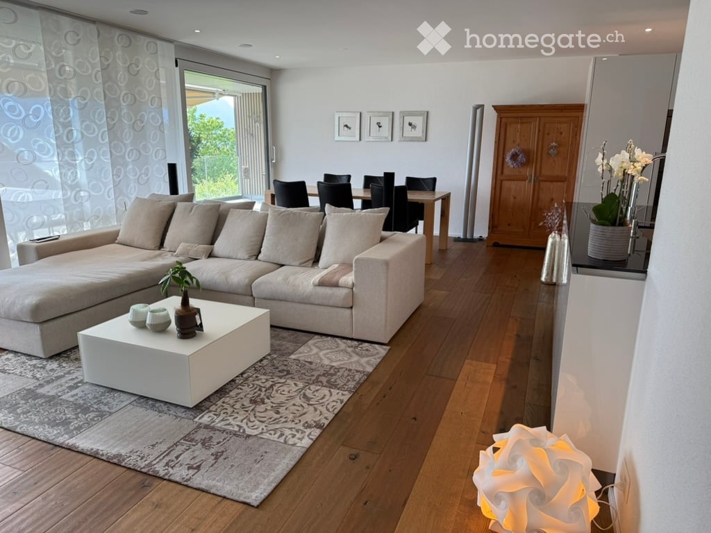 Modern living and dining area with light-colored wood flooring, a large sectional sofa, a white coffee table with a small plant, a dining table with chairs, a wooden cabinet, and decorative vases.