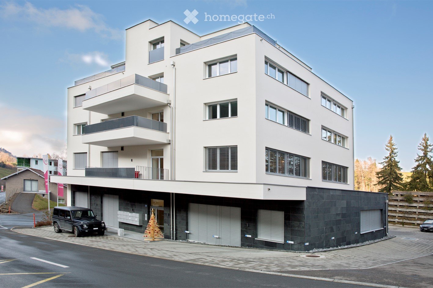 Modern multi-story building with white facade, multiple balconies, and a parking area with a black SUV parked in front.
