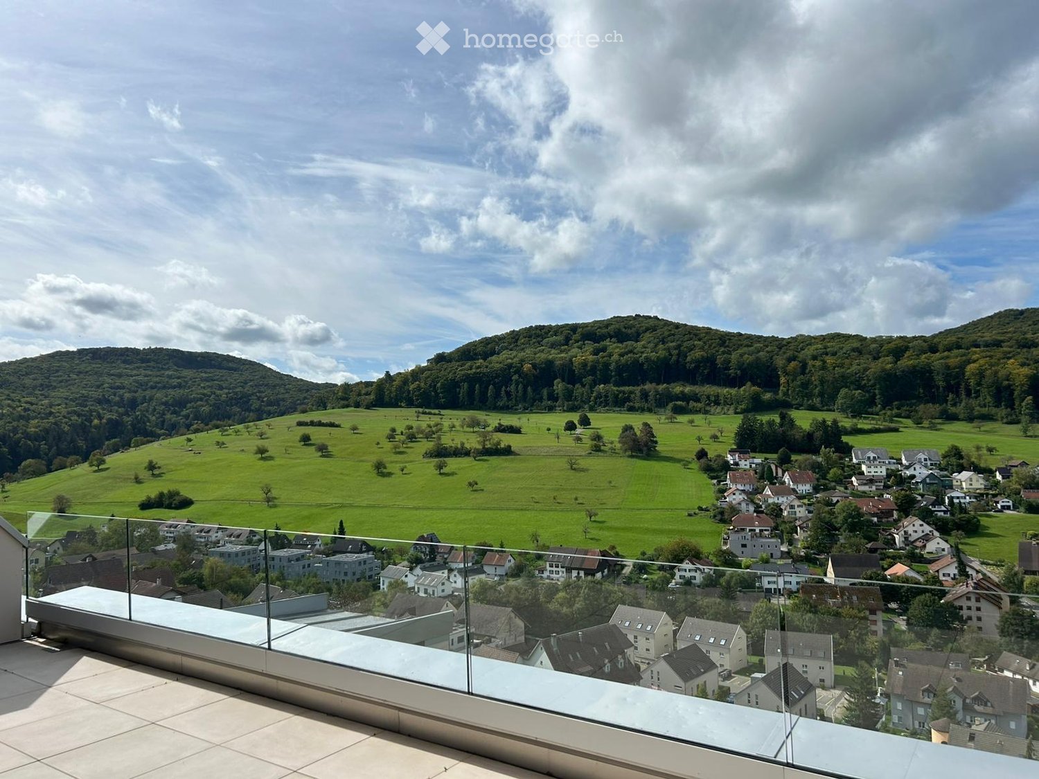 Balcony with a panoramic view of a green valley, scattered houses, and distant hills under a partly cloudy sky.