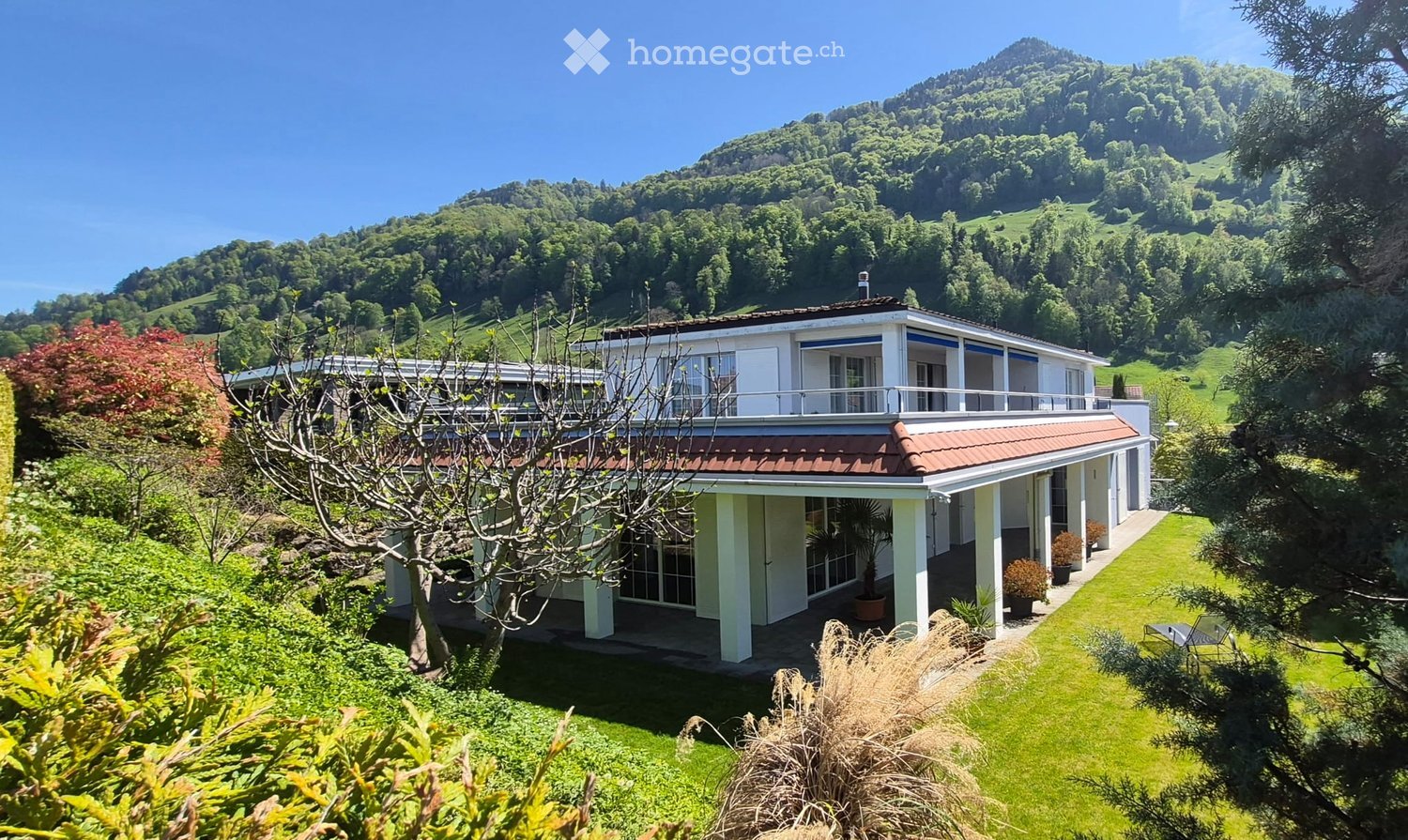 A two-story house with a red-tiled roof and white walls, situated on a raised ground floor with a well-maintained lawn and surrounding greenery, overlooking a scenic mountainous landscape.