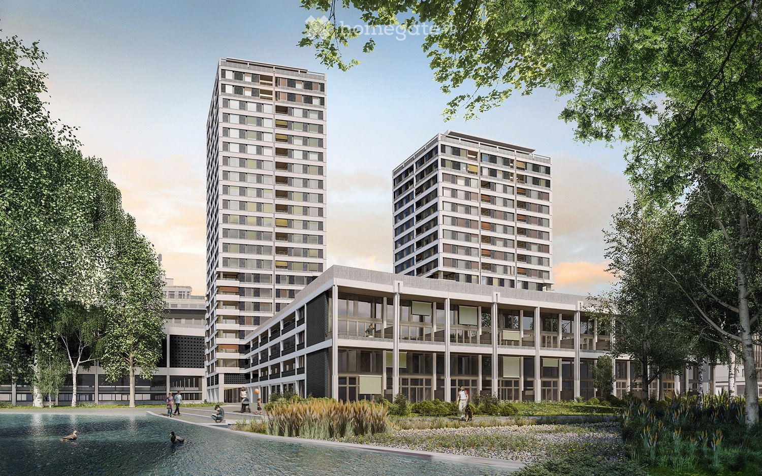 A modern residential complex with two tall apartment buildings and a lower ground-floor building, surrounded by trees and a small water body.
