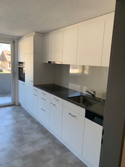 Modern kitchen with white cabinets, black countertops, and stainless steel appliances, including a sink and oven. Large window offers a view of the outdoor area.