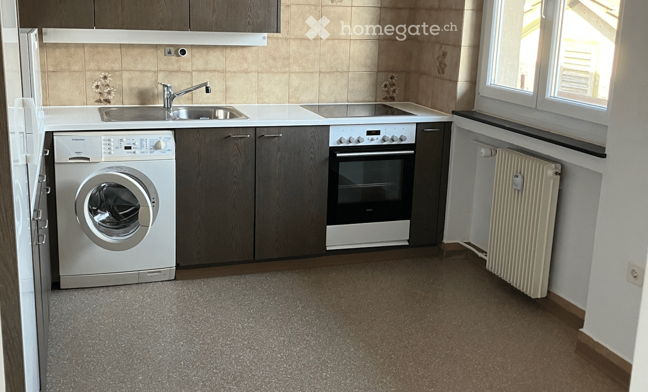 Modern kitchen with a washing machine, oven, sink, faucet, and radiator. The space features wooden cabinets, tiled walls, and a grey carpeted floor.