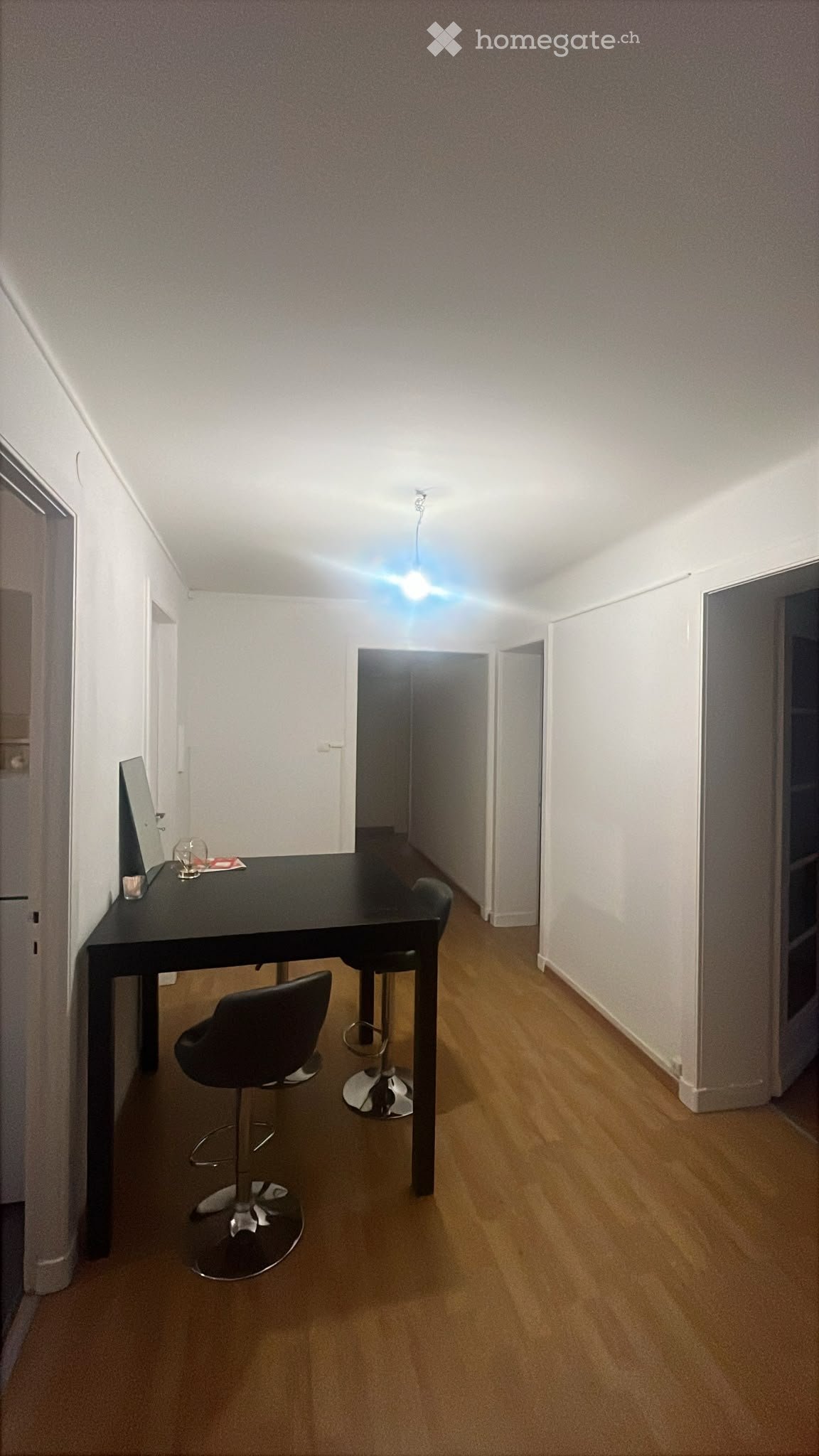 A small, well-lit room with wooden flooring, a black table, two chairs, and two stools. The room has white walls and a ceiling light fixture.