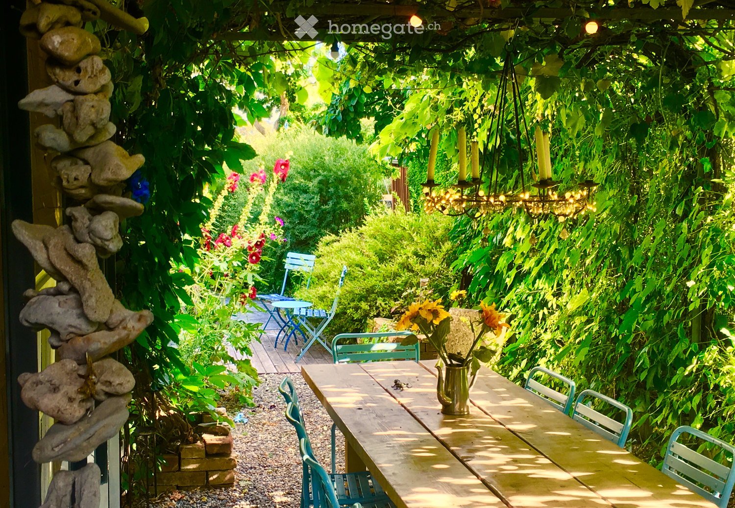 A lush garden area with a wooden table, chairs, hanging chandelier, and colorful flowers, surrounded by greenery and a stone sculpture.