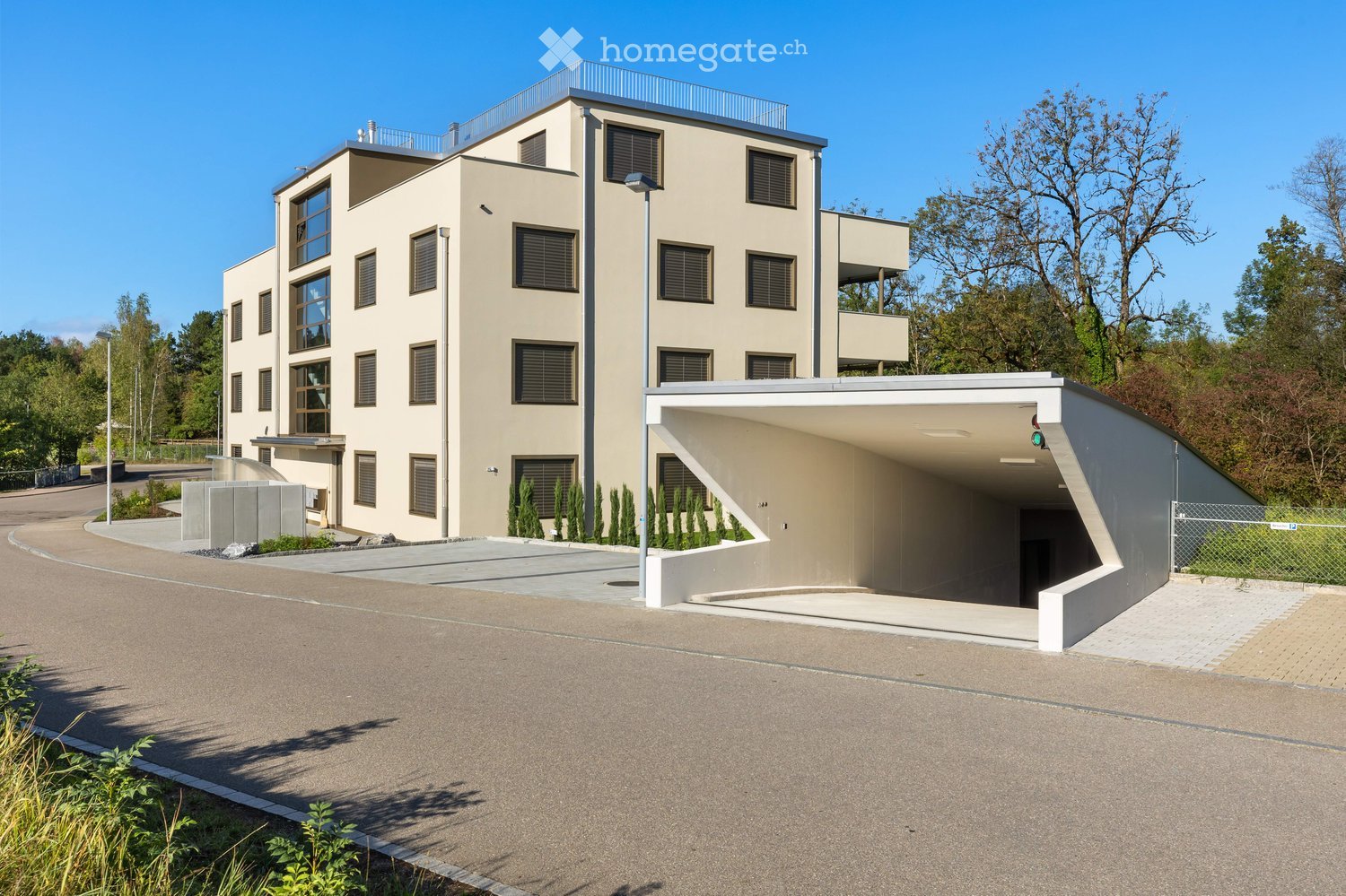 Modern two-story building with beige facade, large windows, and a rooftop terrace. Includes a covered garage area with a traffic light and a fenced perimeter.