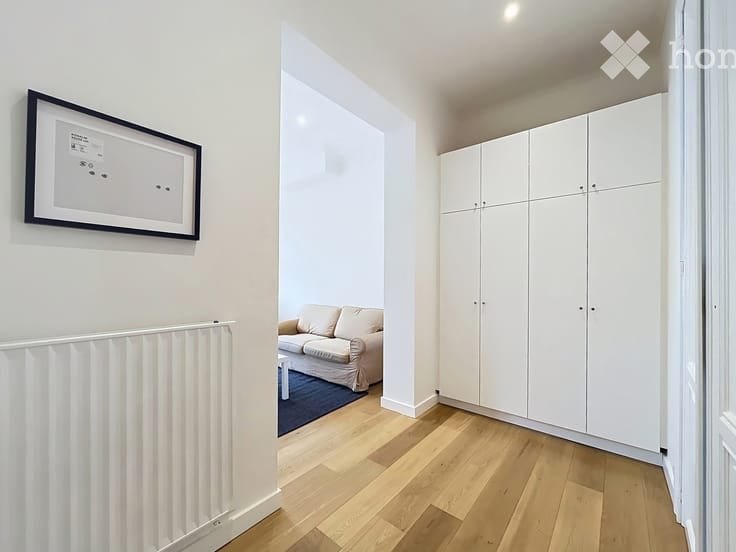 A modern hallway leading into a small living area with a cream sofa, coffee table, and white built-in wardrobes. The hallway features a radiator and a framed picture on the wall.