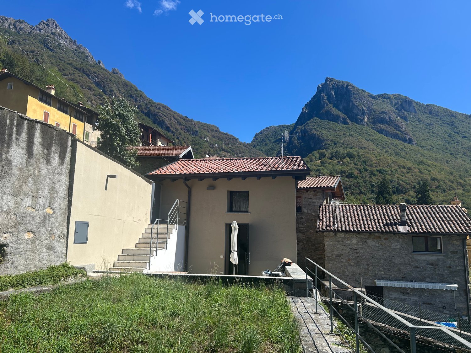 A two-story house with a tiled roof, stone and plaster exterior, and a small porch with an umbrella. The house is situated in a mountainous area with a staircase leading up to the entrance.