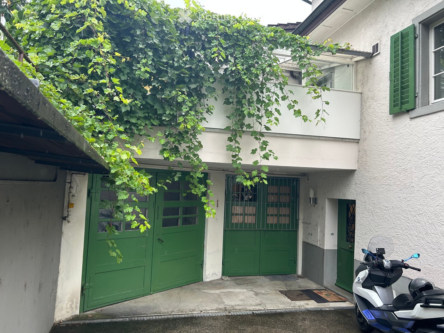 Two-story building with green garage doors, covered by climbing vines, and a scooter parked nearby.