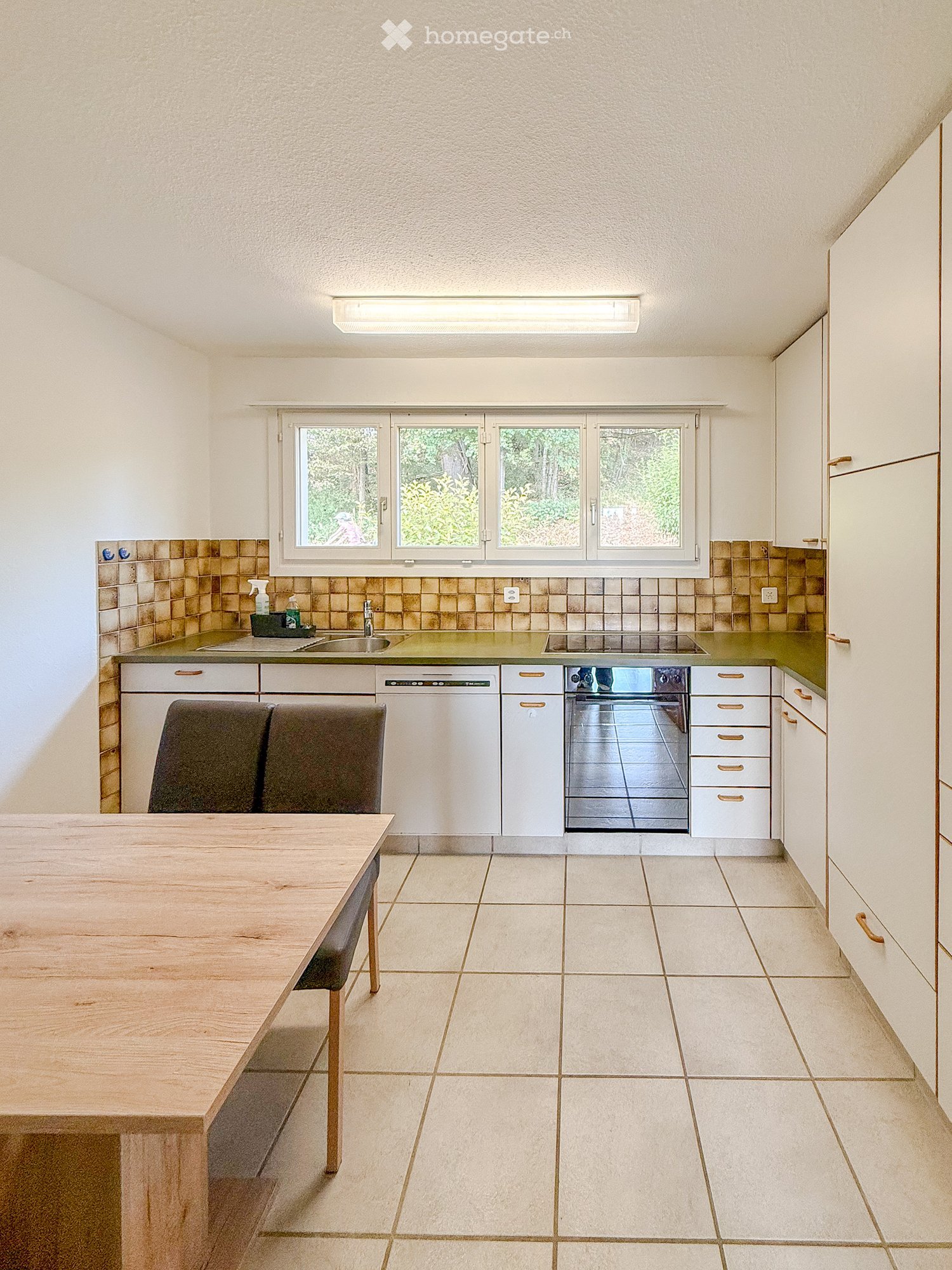 Bright kitchen with white cabinets, a wooden dining table, two chairs, a dishwasher, an oven, and a sink with a faucet. The backsplash features a mosaic tile design, and the countertops are green.