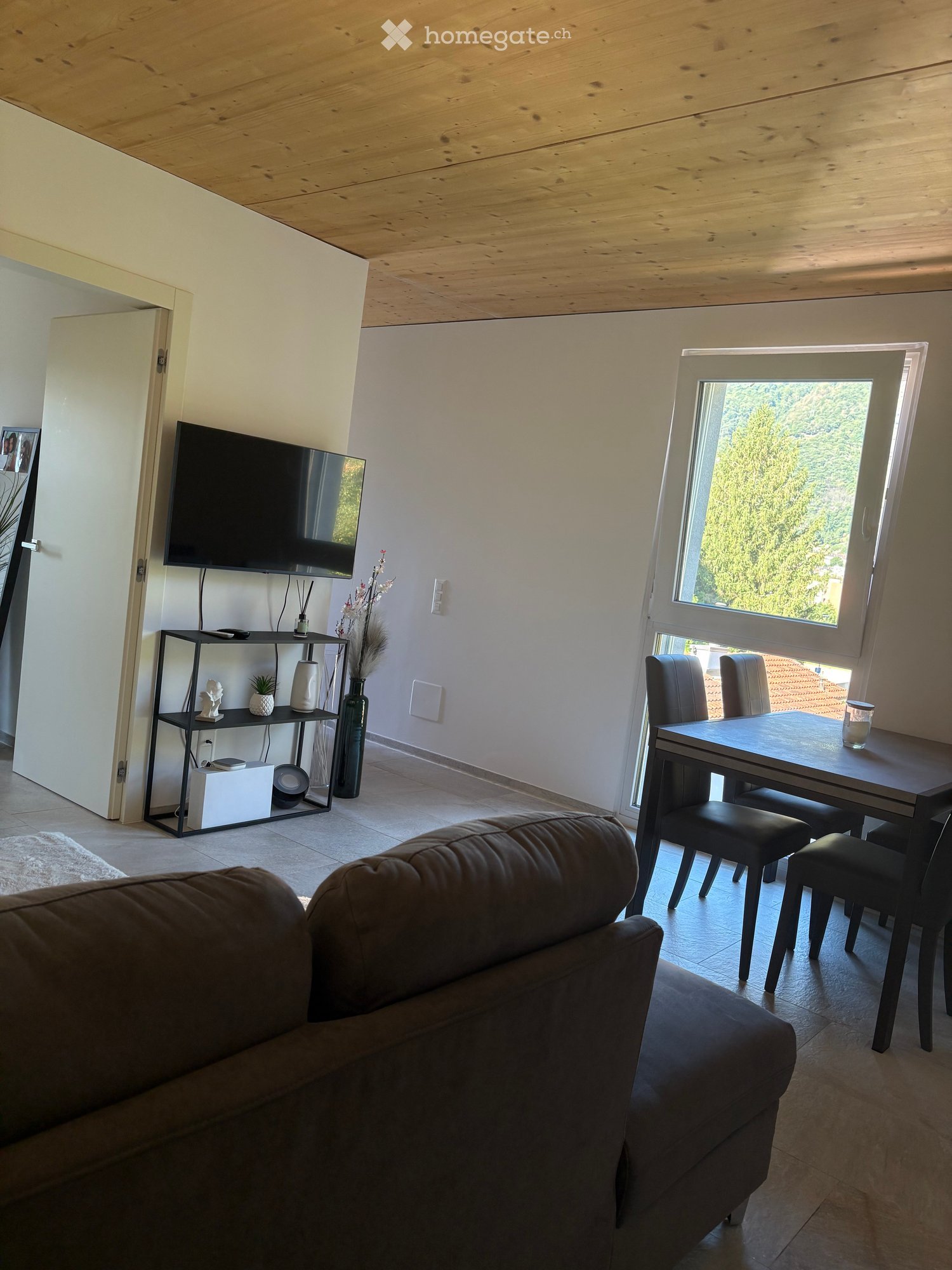 Modern living room with a brown leather sofa, a black dining table with four chairs, a television on a stand, a shelf with decorative items, and a large window with a view of greenery.