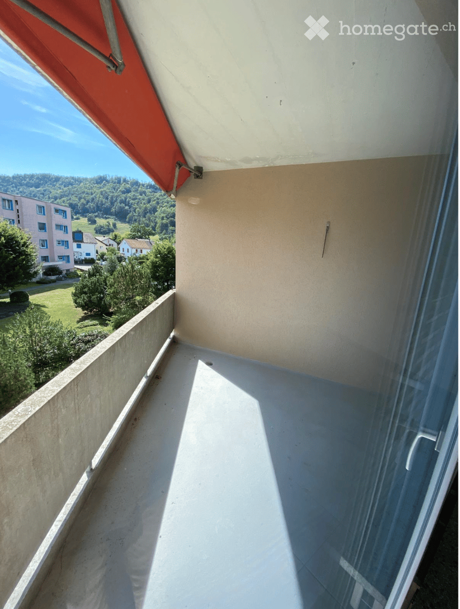 Balcony with a concrete floor, glass railing, and a red awning, overlooking a green landscape with trees and buildings.