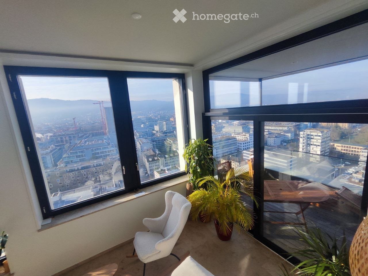 Bright room with large windows offering a cityscape view, furnished with a white chair, a small table, and several potted plants.