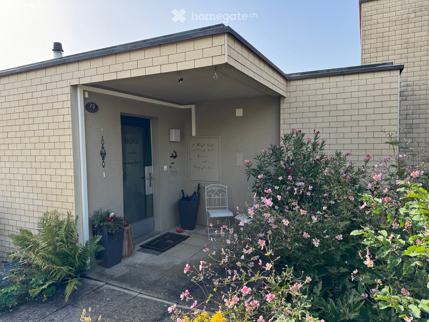 Front entrance of a house with a covered porch, a white metal chair, potted plants, and flowering shrubs.