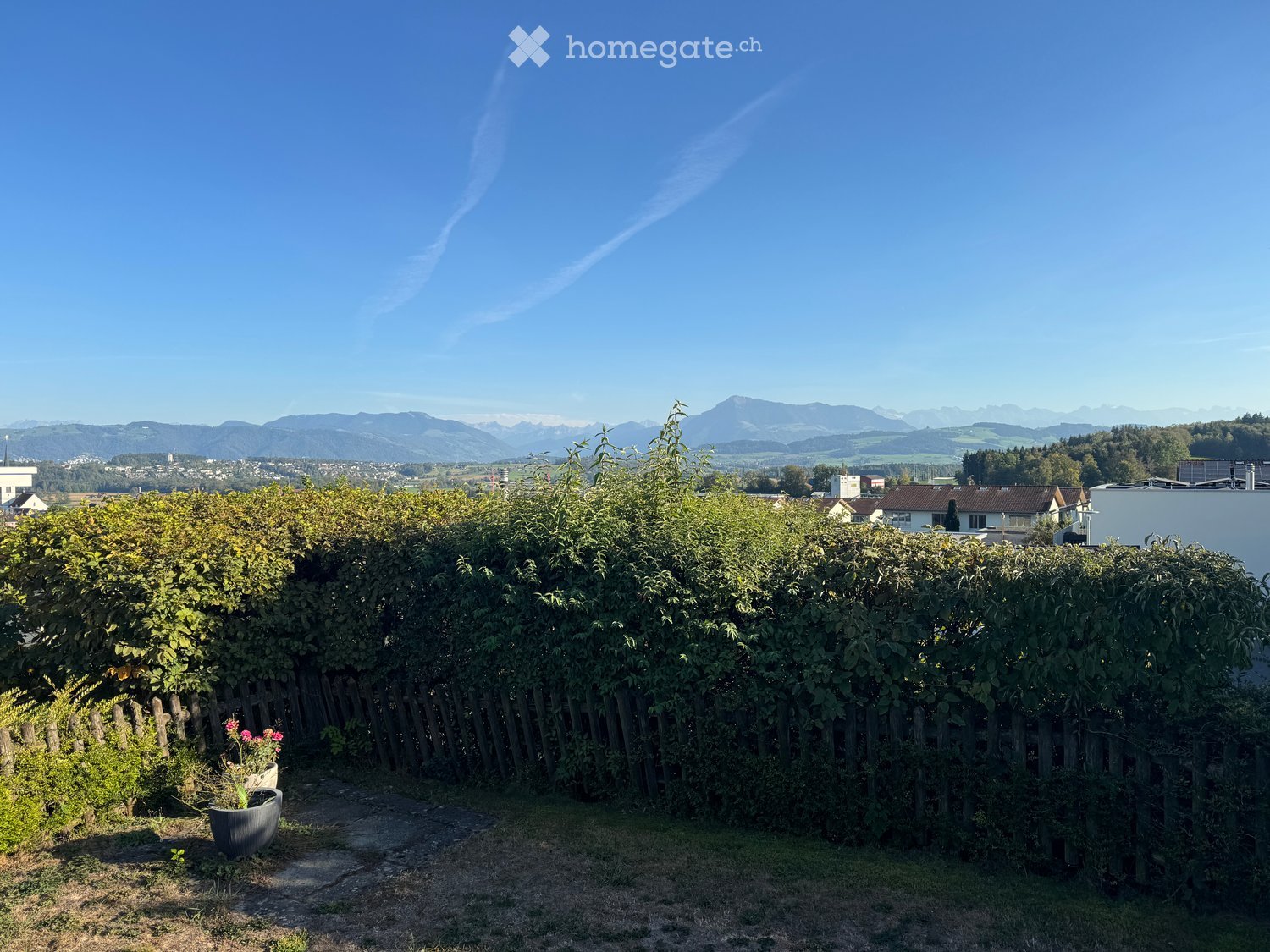 A garden area with a wooden fence, lush greenery, and a view of mountains in the background.