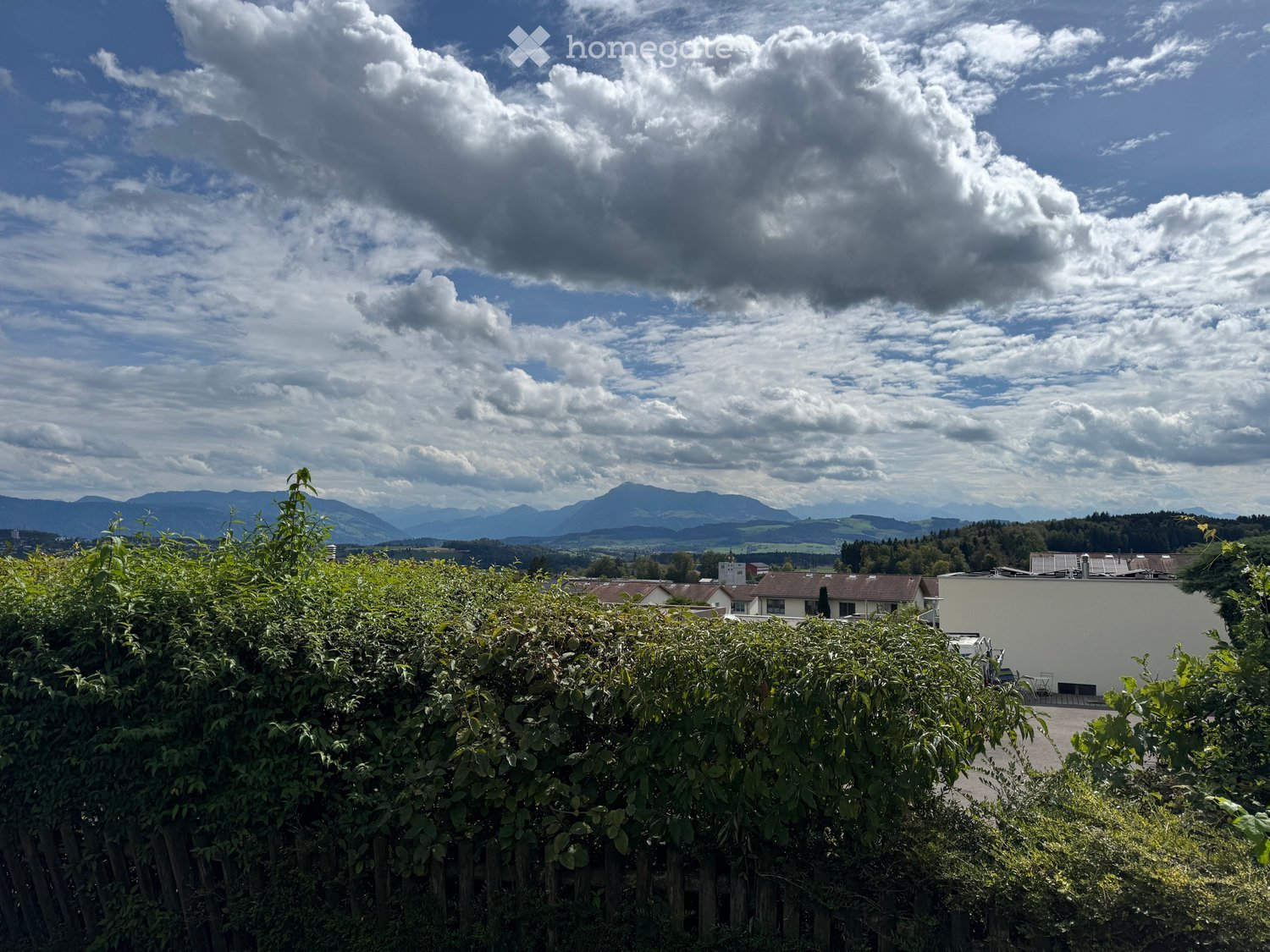 A view from a garden or balcony showing a lush green hedge in the foreground, with a distant view of a town and mountains under a partly cloudy sky.