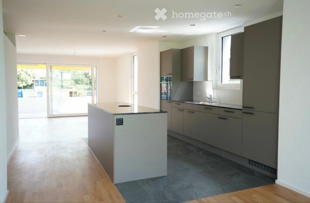 Modern kitchen with light-colored cabinets, a central island, and a dark tiled floor. Features a sink with a modern faucet and ample natural light from large windows and a sliding glass door leading to an outdoor area.