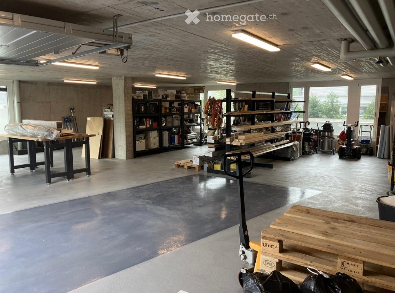 A spacious workshop with concrete floors, exposed ceiling beams, and large windows. The room features a large workbench, metal shelving units filled with tools and supplies, and several pallets and machinery.