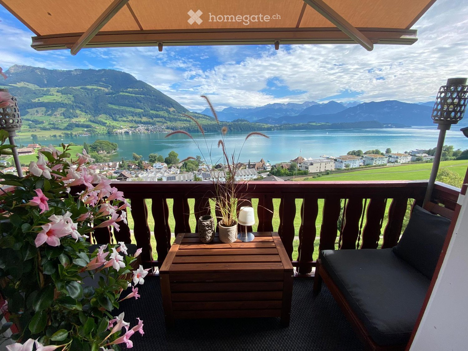 Balcony with wooden railing, overlooking a lake and mountains, featuring a small table, chair, flower pot, and lamp.