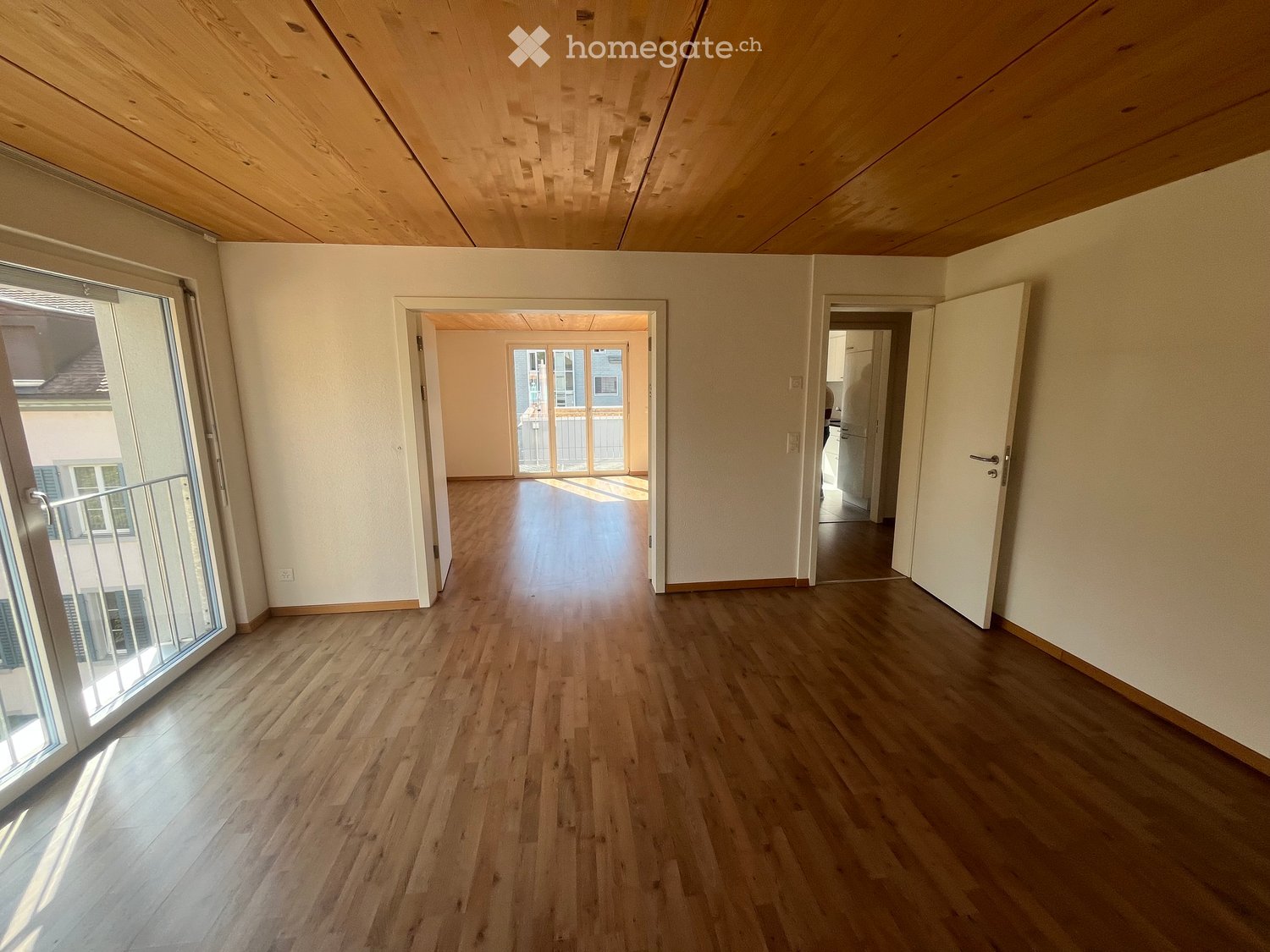 Empty room with wooden flooring, white walls, and a slanted wooden ceiling. Features large windows and open doors leading to other rooms.