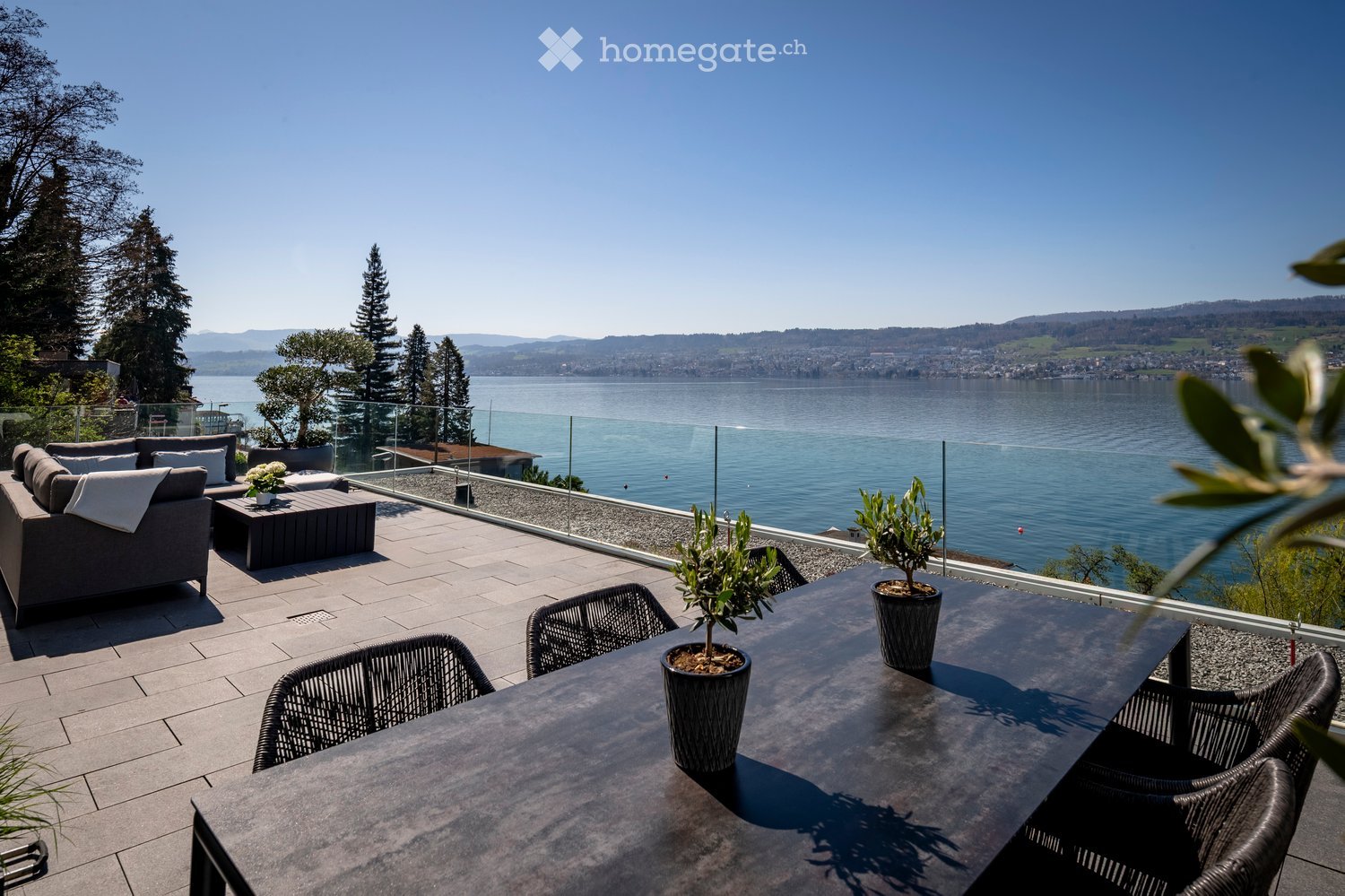 Balcony with a view of a lake, featuring a sofa, coffee table, wicker chairs, and potted plants.
