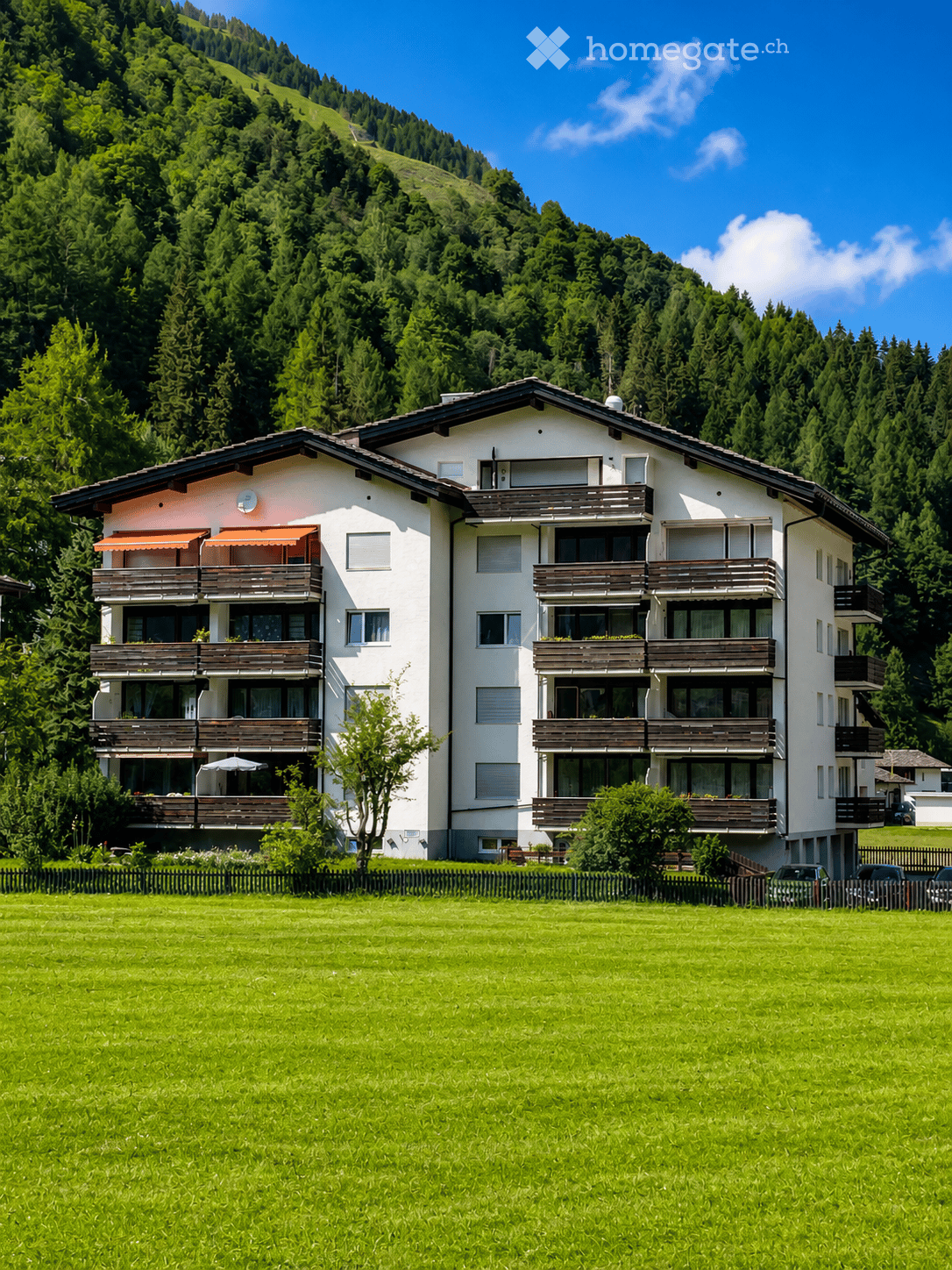 A two-story white building with multiple balconies and awnings, surrounded by a green lawn and trees, with a mountain view in the background.