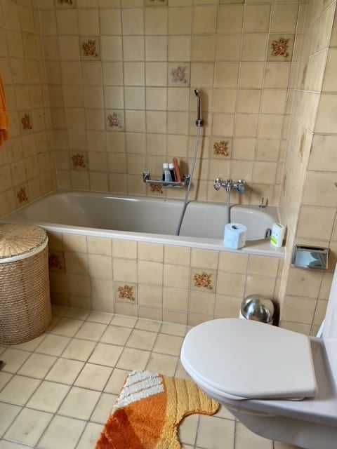 A small bathroom with beige tiled walls and floor, featuring a bathtub, toilet, shower hose, and a wicker basket on the floor.