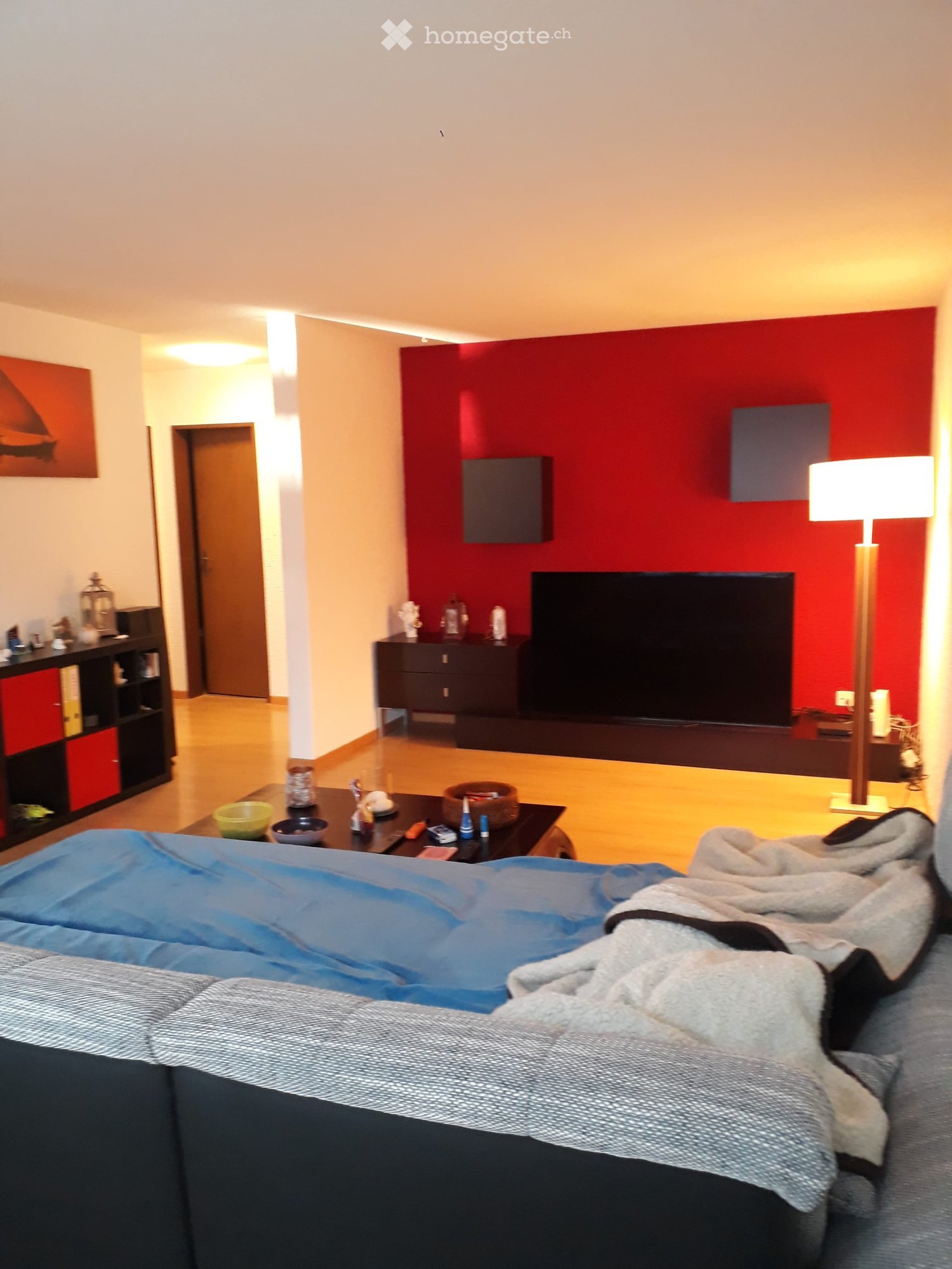 A modern living room with a red accent wall, a large flat-screen TV, a sofa with blankets, a coffee table with various items, a floor lamp, and a shelf with books and decorative items.
