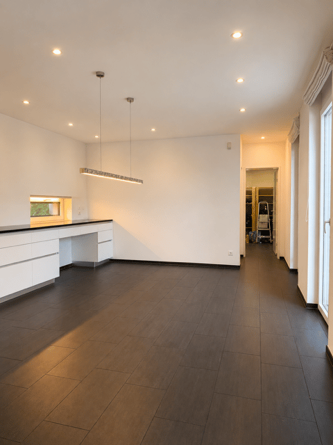 Completely empty room with white walls, dark wooden flooring, and recessed lighting. A small kitchen area is visible with white cabinets and a countertop.