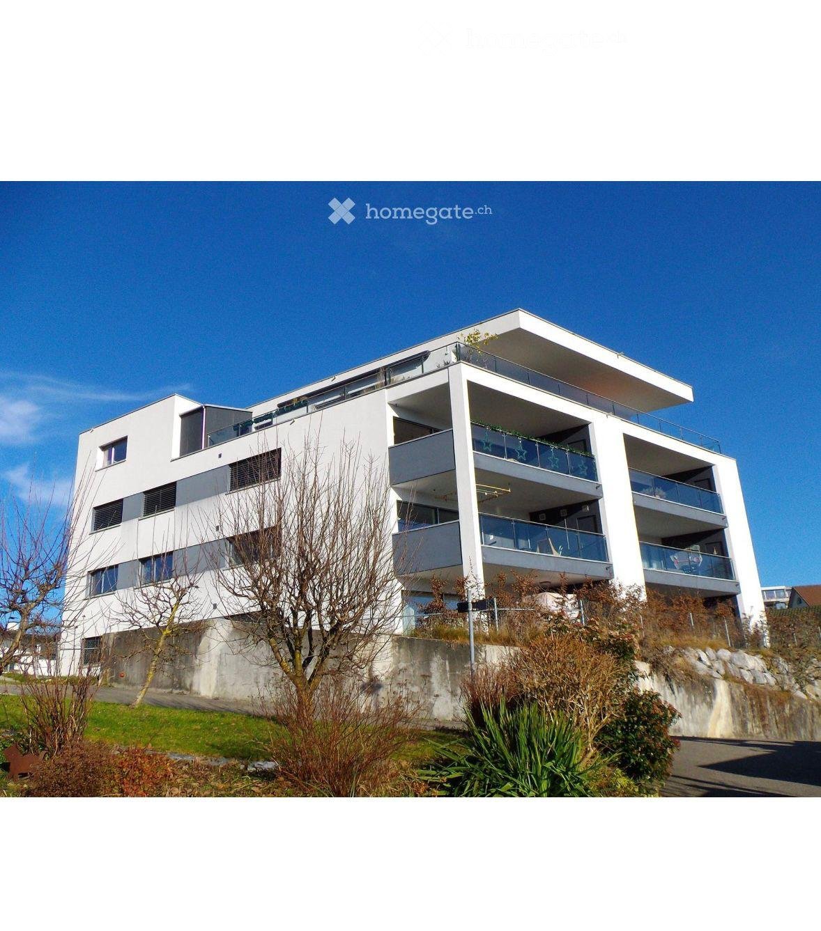 Modern multi-story residential building with white facade, balconies, and contemporary design, situated on a hillside with surrounding greenery.