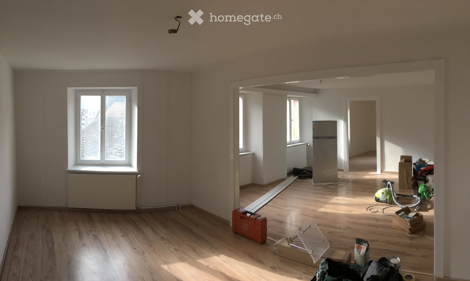 Empty room with wooden flooring, white walls, and large windows. Some construction materials and tools are visible, indicating ongoing renovation.