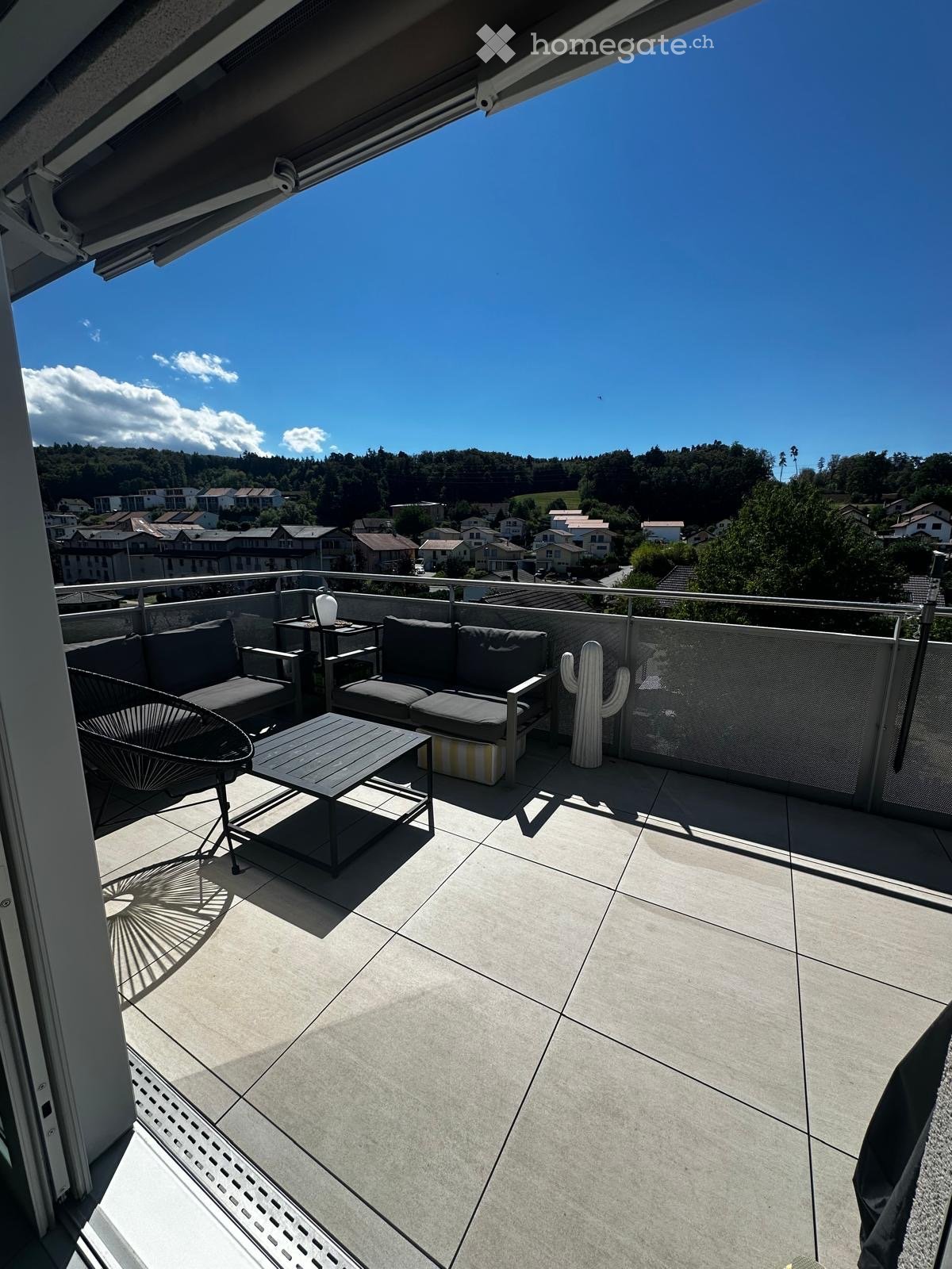 Balcony with a view of the town and surrounding hills, featuring a sofa, table, chair, and a decorative cactus.