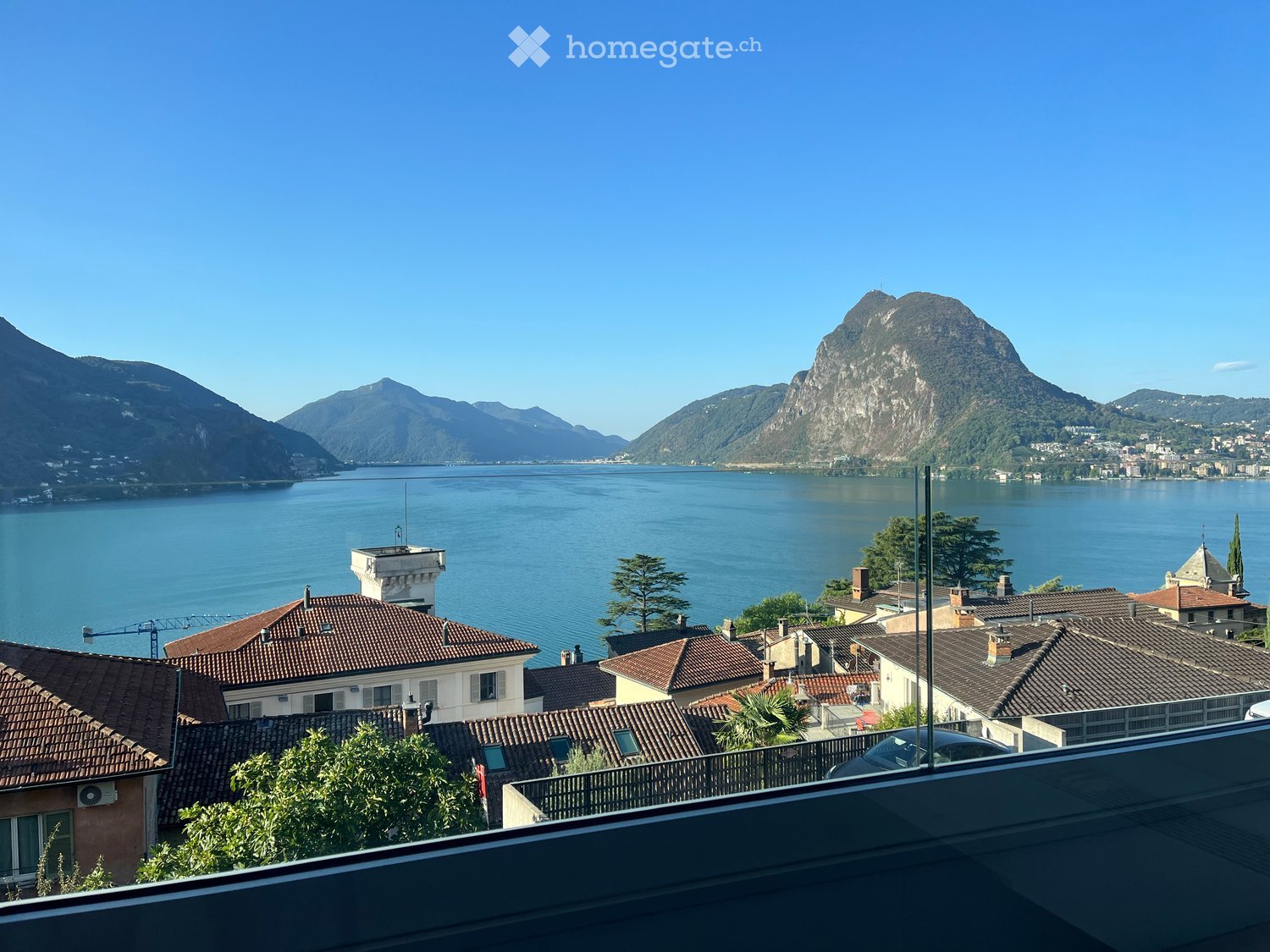Balcony overlooking a scenic lake with surrounding mountains and a town with red-tiled roofs.