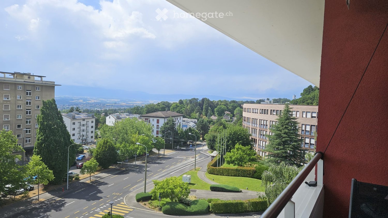 View from a balcony showing a cityscape with buildings, trees, and a road intersection.