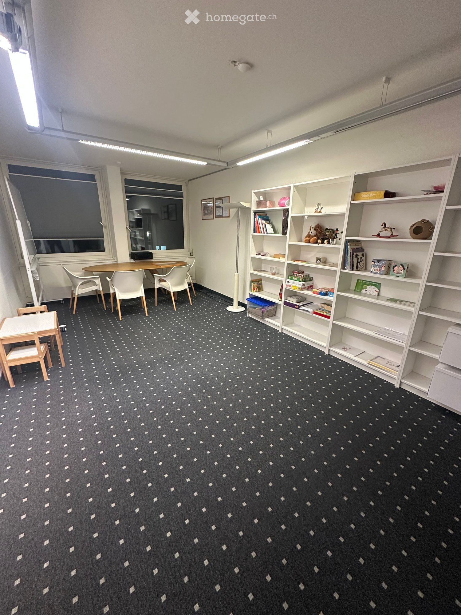 A furnished room with a patterned carpet, a round table with four chairs, a tall floor lamp, and a large bookshelf filled with books and toys.