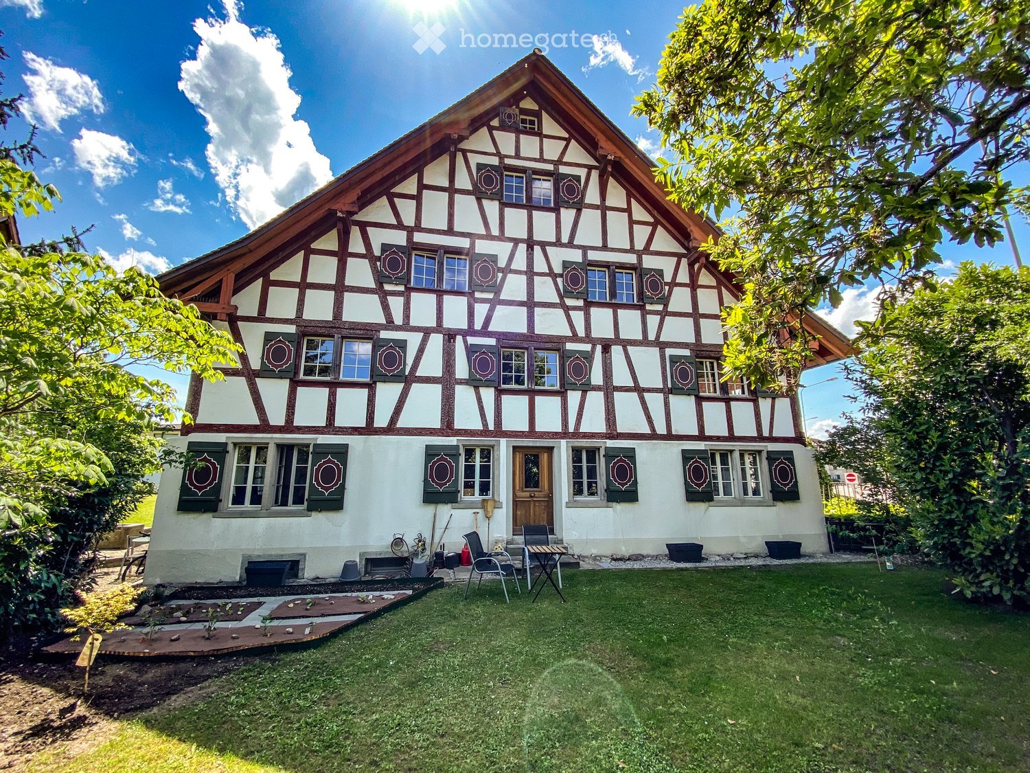 Traditional half-timbered house with white walls, green shutters, and a pitched roof. The front yard features a small patio area with a table and chairs, surrounded by greenery and a well-maintained lawn.