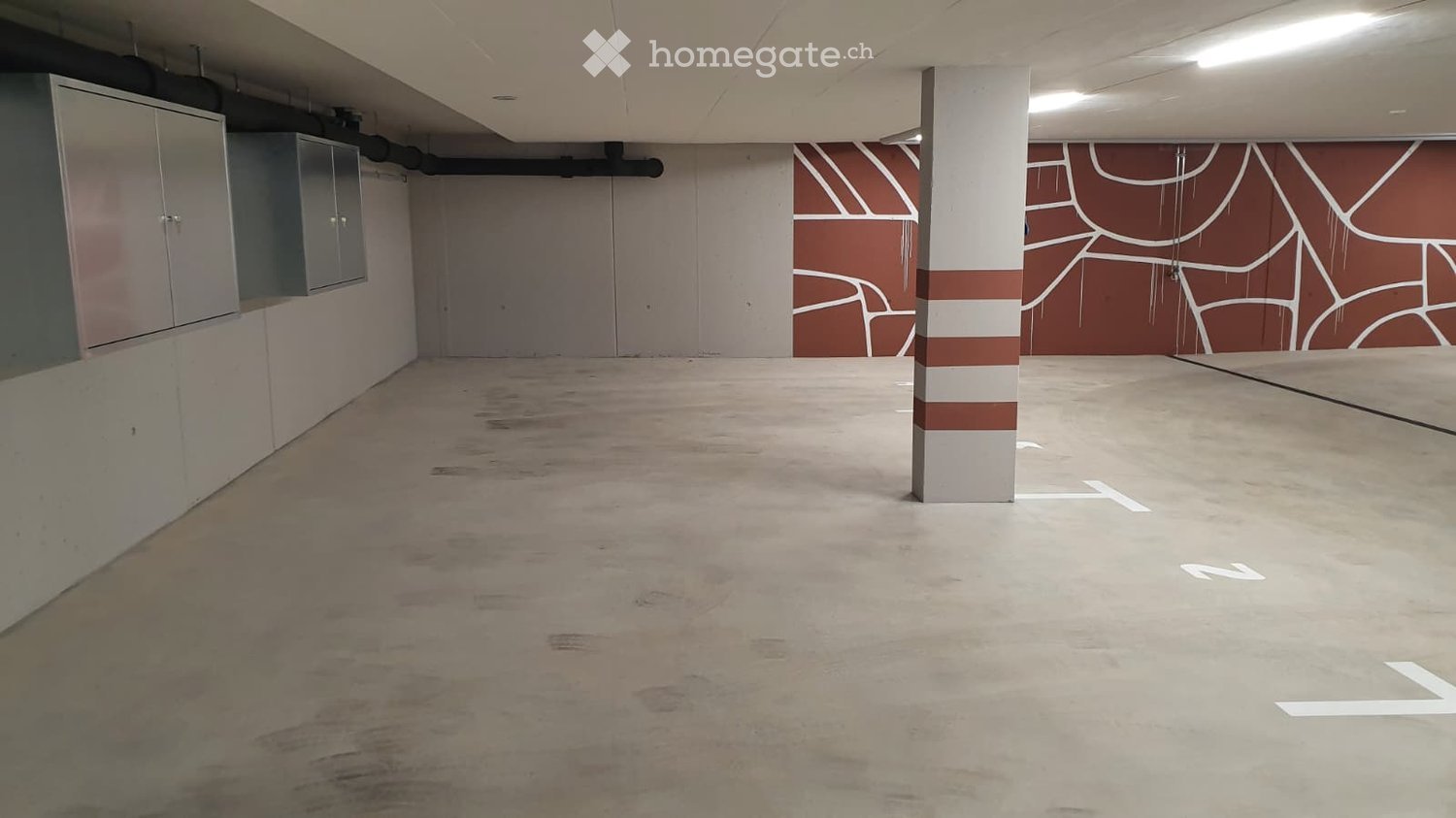 Empty underground parking area with concrete floors, white and red painted walls, and a central support column. No furniture or equipment present.