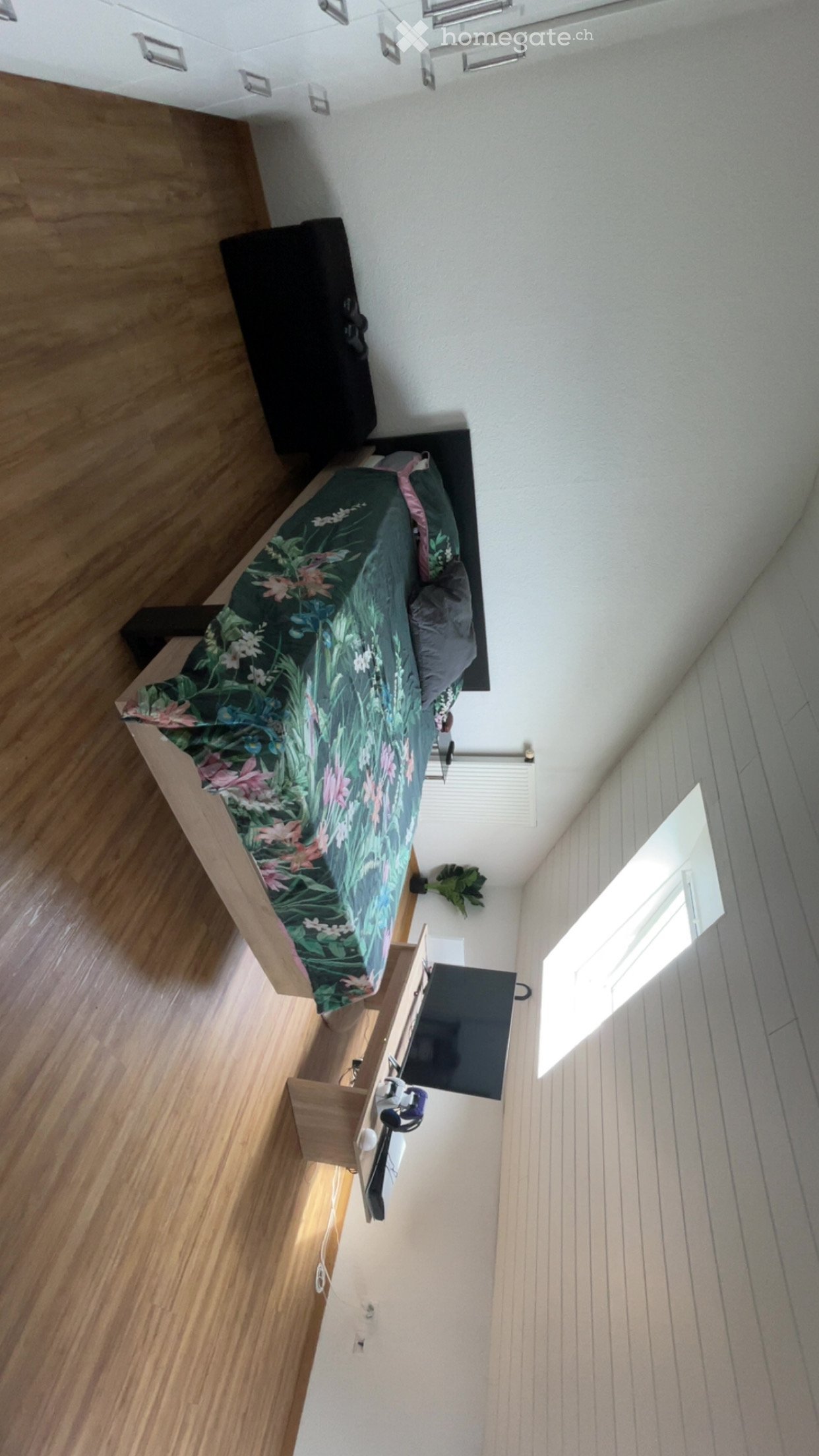 Modern bedroom with wooden flooring, a bed with a green and pink floral duvet, a nightstand with a television, and a small plant near the window.
